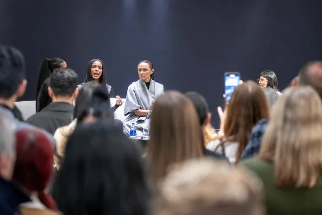 A Sisters founder on stage mid-sentence beside a guest, seen over the heads of the audience